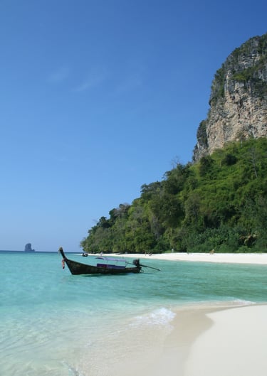 A traditional Thai longtail boat floats in turquoise water by a white sand tropical beach and limestone cliffs.