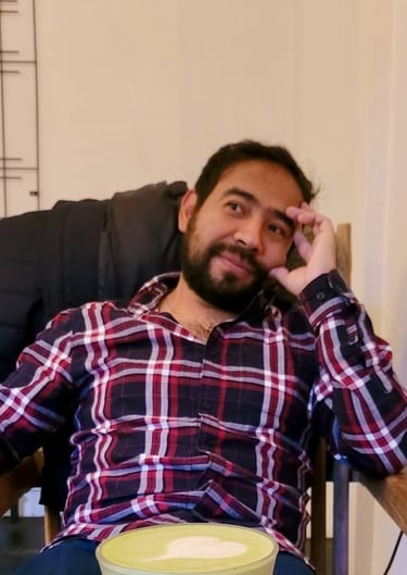 Tsinvari sitting in a chair, wearing a plaid shirt, with his hand near his face in a thoughtful pose