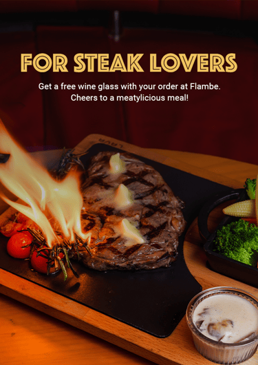 Flaming steak served on a wooden board with grilled vegetables and sauce. Text promotes a free wine.