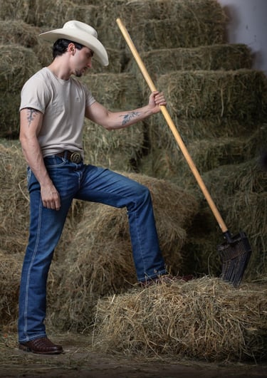 A ranch hand in a white cowboy hat and blue jeans holding a pitchfork in front of hay bales.