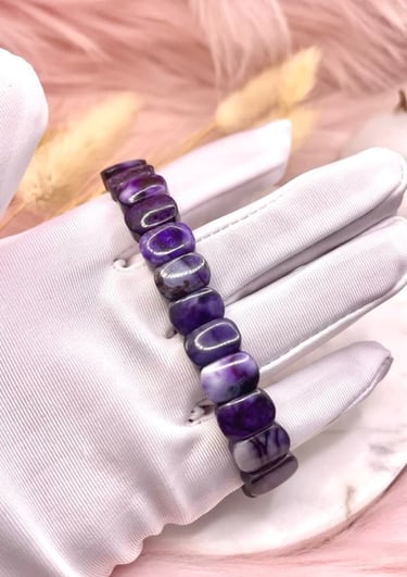 A gloved hand holding a deep purple beaded bracelet against a soft, neutral-toned background.