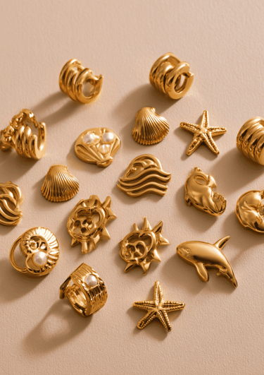 Colección de dijes y anillos dorados de acero inoxidable con formas marinas sobre fondo beige.