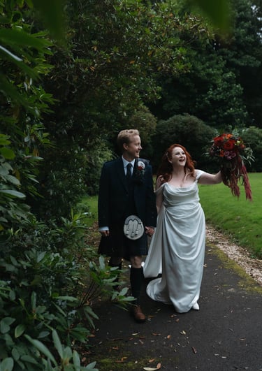 A bride in a white wedding gown and groom in a traditional Scottish kilt walk through a lush garden path.