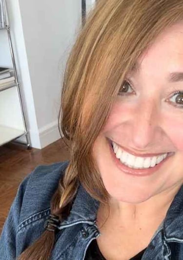 Smiling woman with reddish-brown hair in a side braid wearing a denim jacket in a brightly lit room.