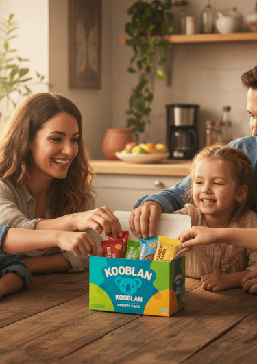 A smiling family gathered around a kitchen table sharing an open variety box of Kooblan snack bars, 
