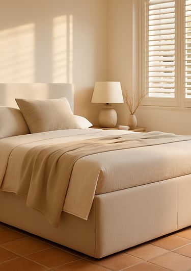 Cama con canapé abatible y ropa de cama beige en dormitorio mediterráneo con ventanas y luz suave.