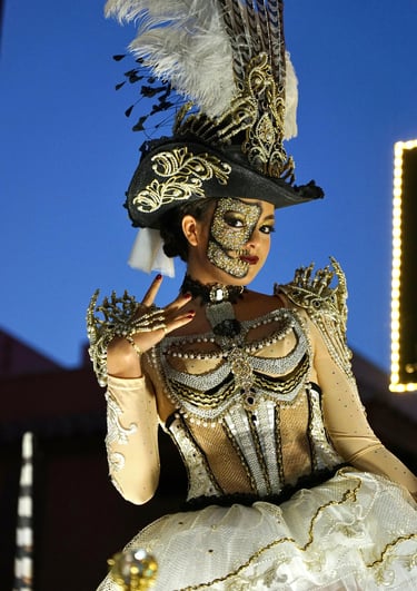 Woman in an ornate gold and white carnival costume with a skull face mask and feathered hat.