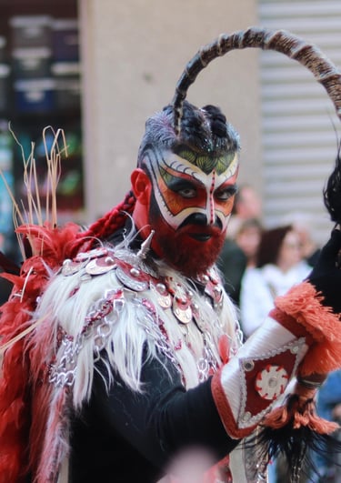 Man in elaborate red and white tribal costume with face paint and horns at a festival.