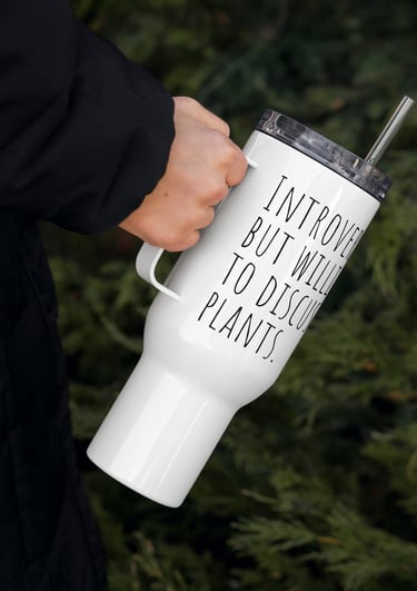 Hand holding white plant-themed travel mug outdoors near green foliage.