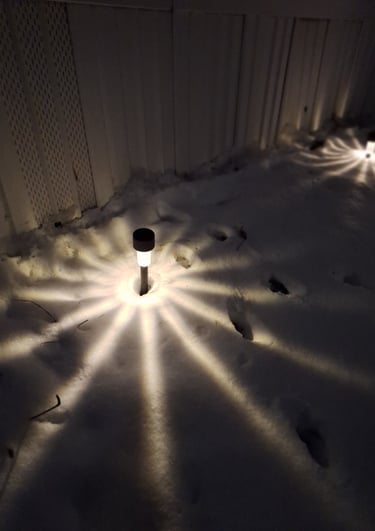 Solar pathway lights casting a starburst pattern onto white snow along a garden fence at night.