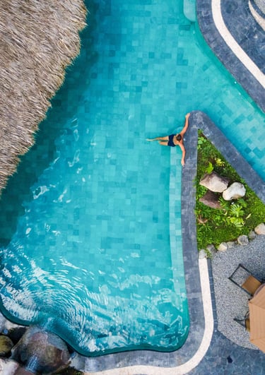 Aerial view of a woman relaxing in a luxury tropical resort swimming pool next to a thatched roof hut.