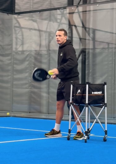A man practicing padel on a blue indoor court with a racket and a Head ball cart.
