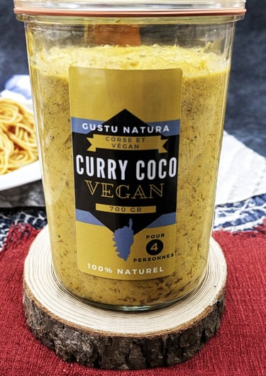 Glass jar of Gustu Natura vegan curry coco sauce on a rustic wooden coaster with pasta background.