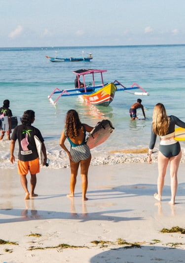 Surfers walking on a white sand beach in Lombok with a colorful outrigger boat in the ocean.