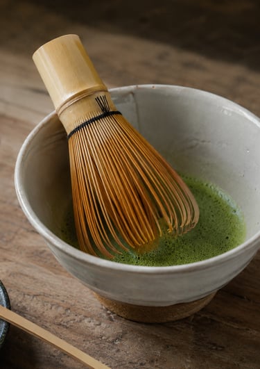 Traditional bamboo whisk stirring frothy green matcha tea in a ceramic bowl on a wooden surface.