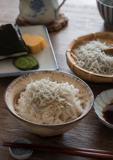 A traditional Japanese breakfast with a bowl of shirasu whitebait over steamed rice, onigiri, and tamagoyaki.