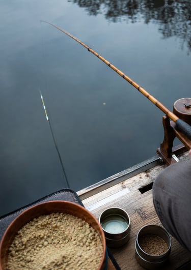A fisherman holds a bamboo fishing rod over calm lake water with bait bowls nearby on a wooden dock.
