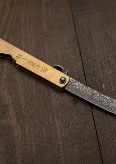 A Japanese Higonokami folding pocket knife with a Damascus steel blade and brass handle on wood.