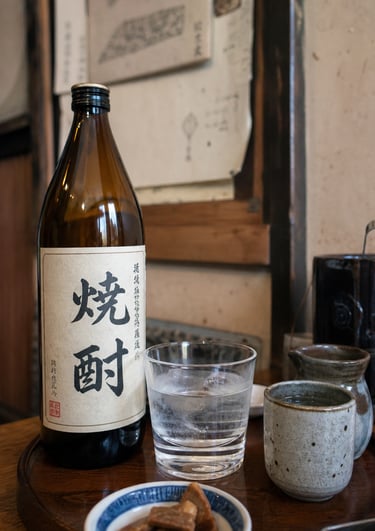 Japanese shochu bottle and glass on a wooden tray with traditional ceramic sake cups and appetizers.