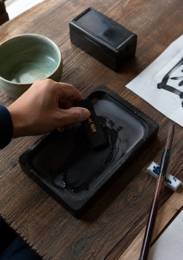A hand grinding an inkstick on a stone inkwell for traditional Chinese calligraphy.