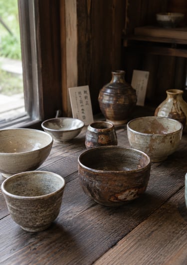 Handcrafted Japanese ceramic tea bowls and pottery displayed on a rustic wooden table by a window.