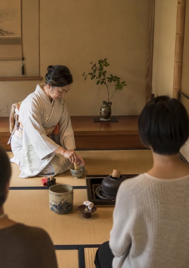 A Japanese woman in a kimono performs a traditional matcha tea ceremony for guests on tatami mats.