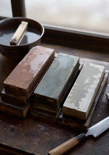 A collection of wet Japanese natural whetstones and a chef knife on a wooden workbench for professional sharpening.