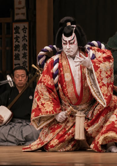 A Kabuki performer in a red floral kimono with dramatic makeup and traditional musicians on a Japanese stage.