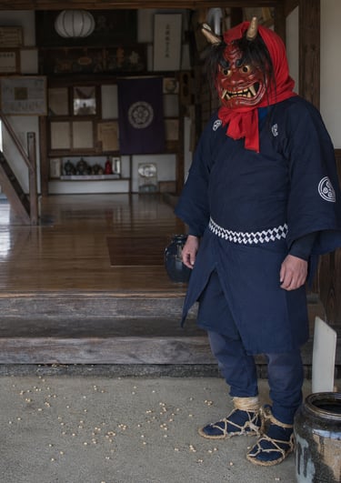 A man wearing a traditional red Japanese Oni demon mask and blue kimono at a temple entrance.