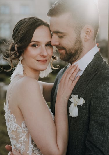 die Braut im romantischen Boho-Look, hat eine Hand auf den Bräutigam gelegt, der sie liebevoll ansieht und eng an sich zieht