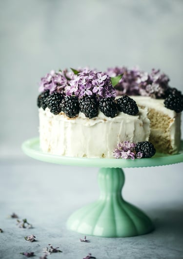 Pastel de vainilla en capas con betún blanco, decorado con moras frescas y flores lilas moradas.