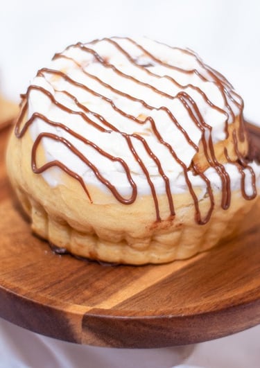 Rollo de canela recién horneado con glaseado blanco y un chorrito de chocolate sobre un pedestal