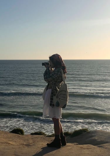 Vibey full body candid shot of me holding camera up to my face, standing on cliffs over ocean.Earthy