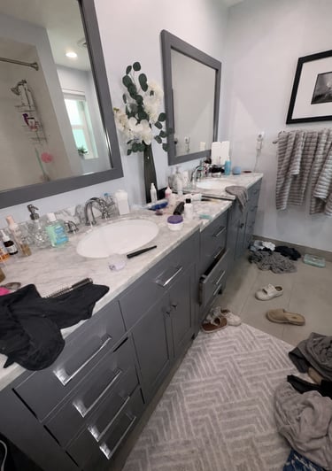 Messy modern bathroom with dual gray vanities, marble countertops, and clothes on the floor.