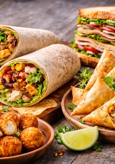 A variety of party food including chicken wraps, club sandwiches, golden samosas, and crispy potato bites.