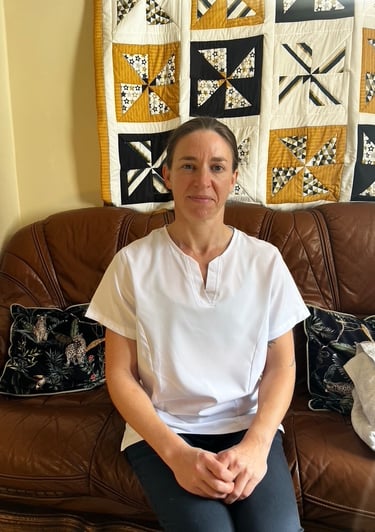 Image shows Rae sitting on a brown leather sofa in a white masseuses top and blue chinos.