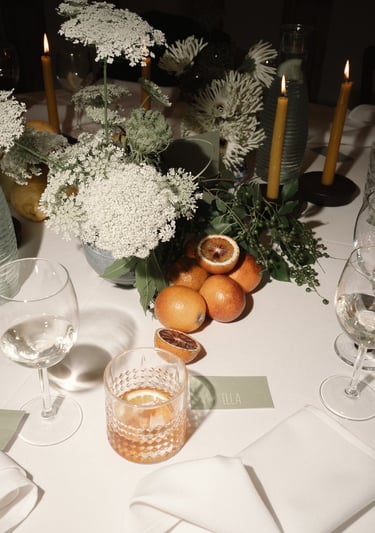 Elegantiškai dekoruotas stalas su baltomis gėlėmis, žalumynais, raudonais apelsinais ir kokteiliu