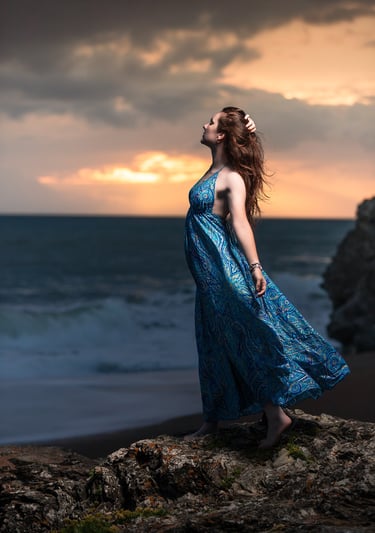 Creative portrait of a woman in a blue dress standing on rocks at sunset