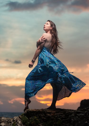 Portrait créatif d’une femme en robe bleue posant sur des rochers au coucher du soleil.