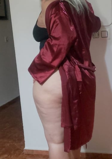 Escort madura colombiana en bata roja de satén, de rodillas sobre el sofá, mostrando sus piernas provocativas.