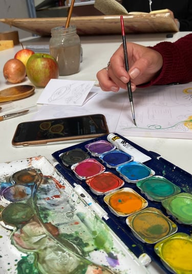 Artist painting a watercolor autumn still life with a pumpkin, apples, and watercolor palette.