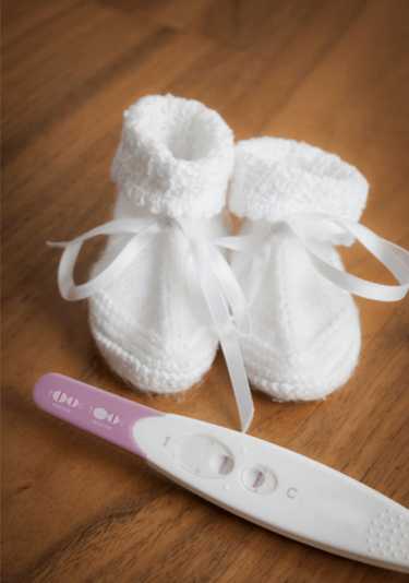 Positive pregnancy test result next to white knitted baby booties on a wooden background.
