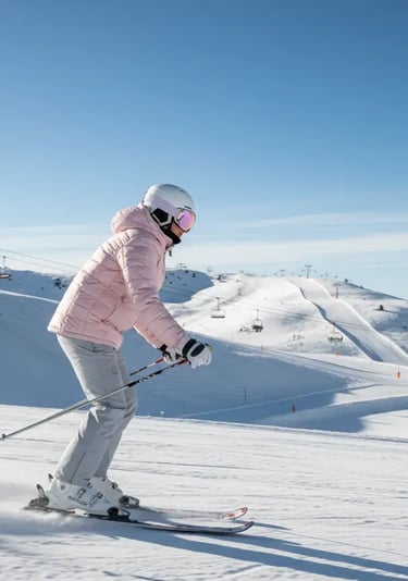 A woman skiing down a snowy slope in a light pink puffer jacket and grey ski pants, showcasing a fem