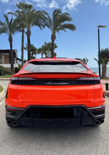 Rear view of a bright orange Lamborghini Urus luxury SUV parked by palm trees near the ocean.