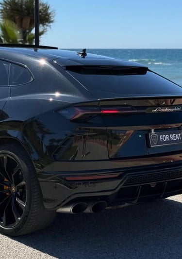 Black Lamborghini Urus luxury SUV available for rent parked on a coastal road by the ocean.