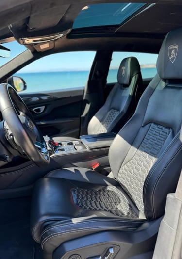 Luxury interior of a Lamborghini Urus with black leather seats and honeycomb stitching parked by the ocean.