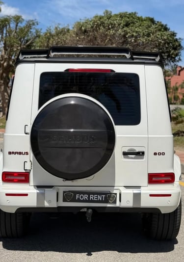 White Brabus 800 luxury SUV for rent parked on a sunny street with carbon fiber spare tire cover.