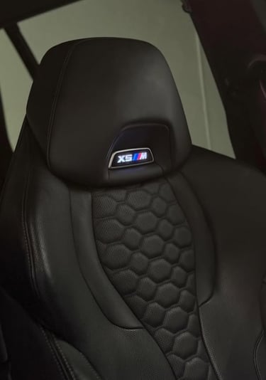Close-up of a black leather BMW X5M sport seat with quilted hexagonal pattern and illuminated M badge logo.