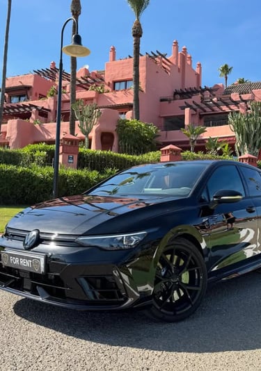 Black Volkswagen Golf R for rent parked in front of a luxury pink resort with palm trees.