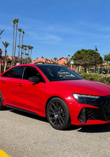 A vibrant red Audi RS3 luxury sedan parked on a sunny resort road with palm trees and blue sky.